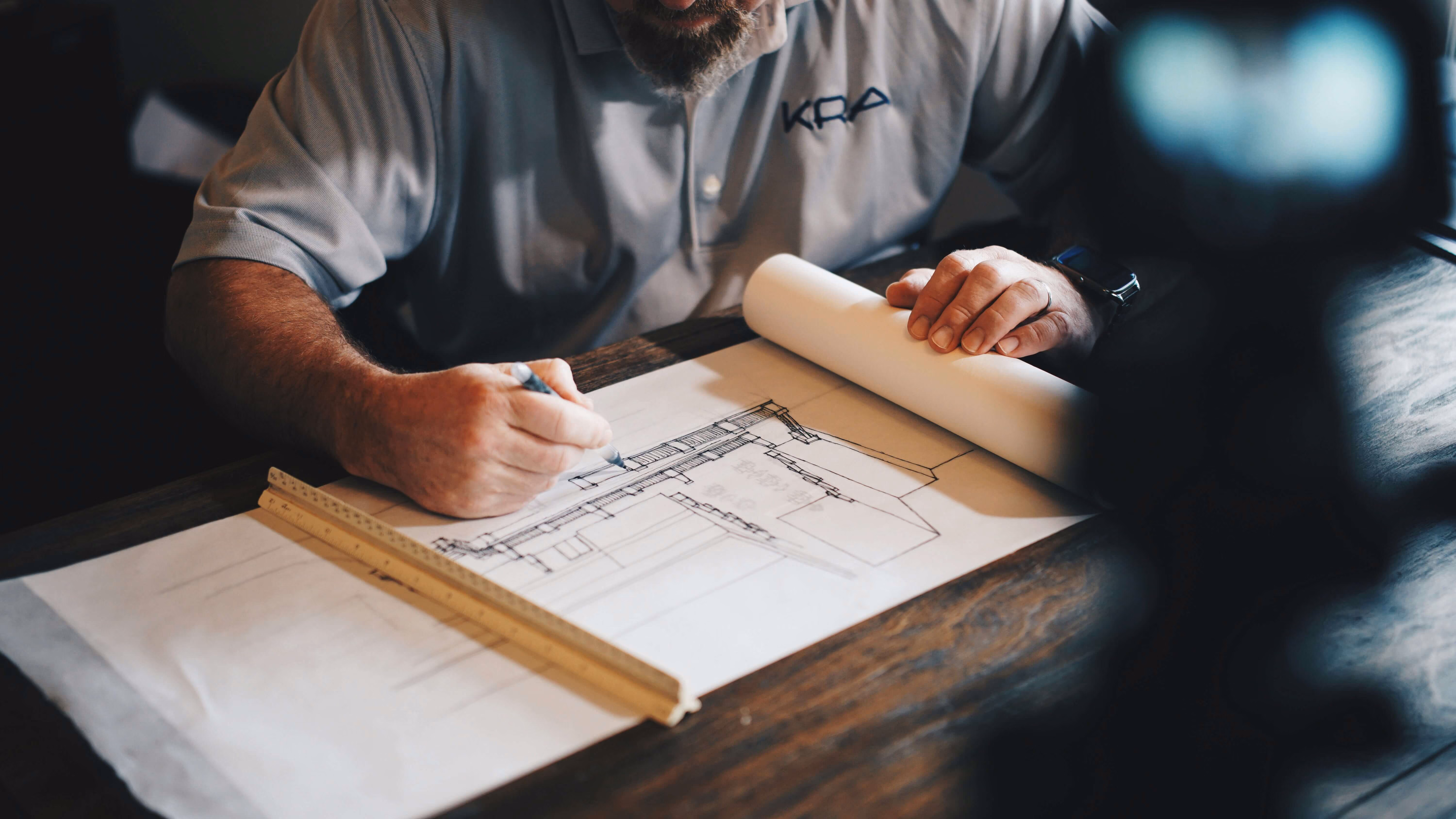 Mann in grauem Hemd zeichnet einen Architekturplan auf einem Holztisch mit Lineal.