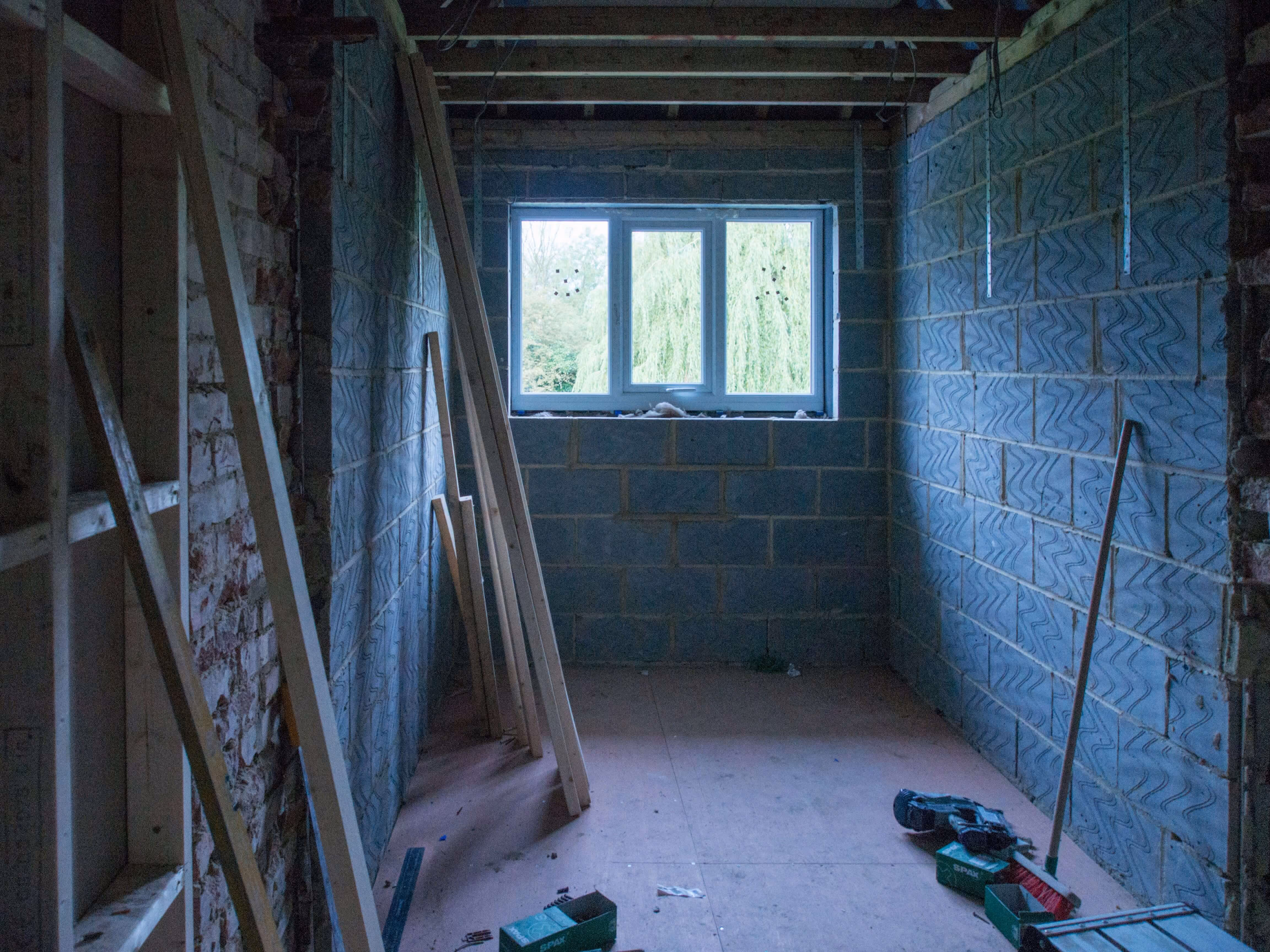 Unfertiger Raum mit blauen Mauersteinen, Holzbalken an der Decke, einem Fenster und Bauholz sowie Werkzeug auf dem Boden.